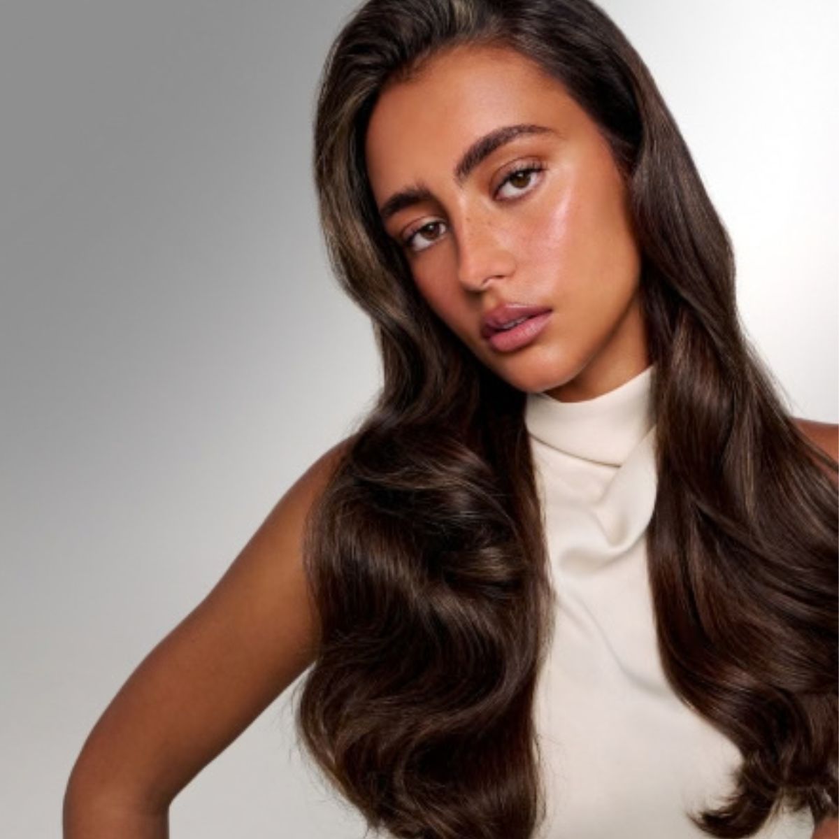 Femme aux longs cheveux châtain foncé ondulés portant un haut crème sans manches, sur fond gris pâle avec une touche de bleu