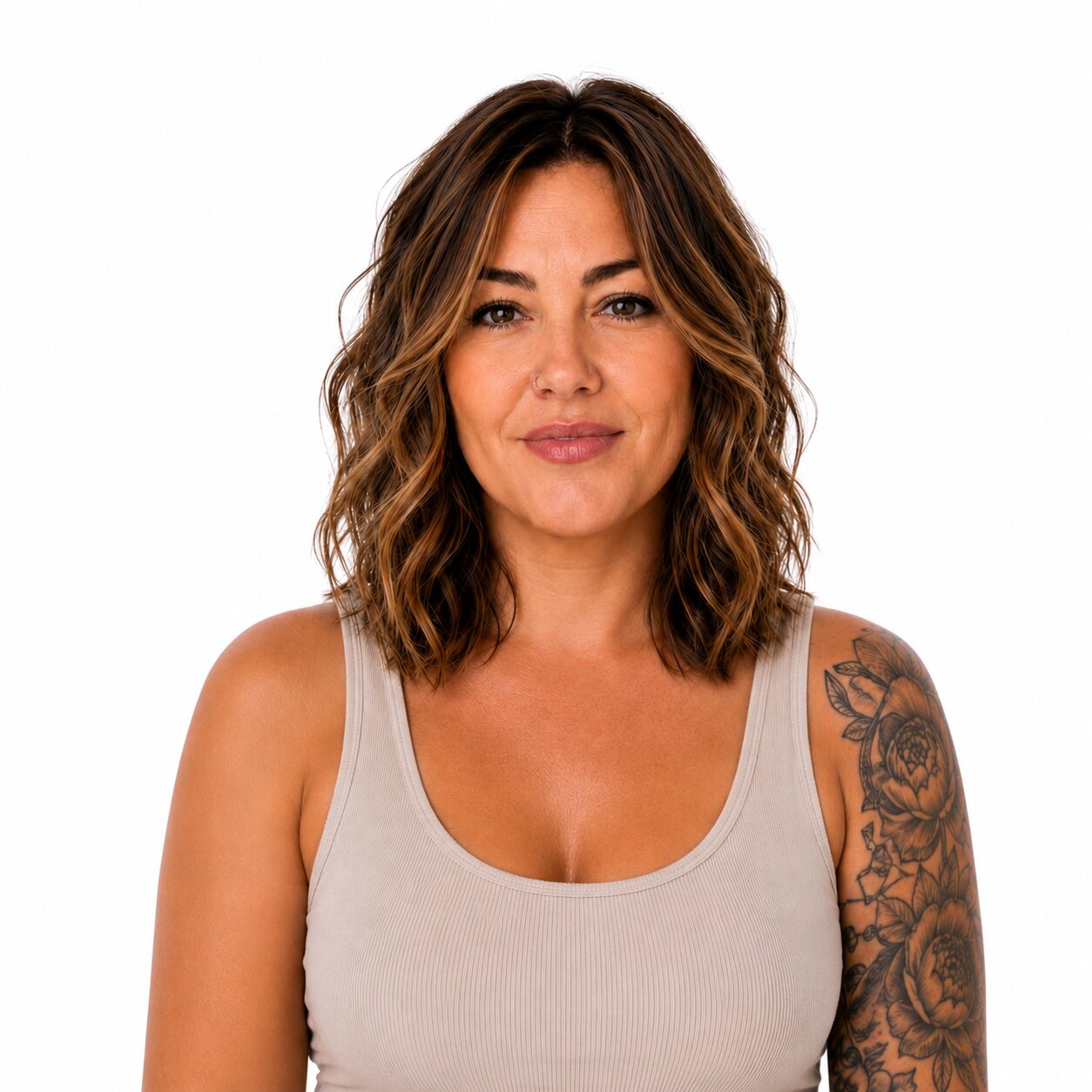 A woman with shoulder-length brown hair and a tattoo on her left arm, wearing a beige ribbed tank top against a plain white background.