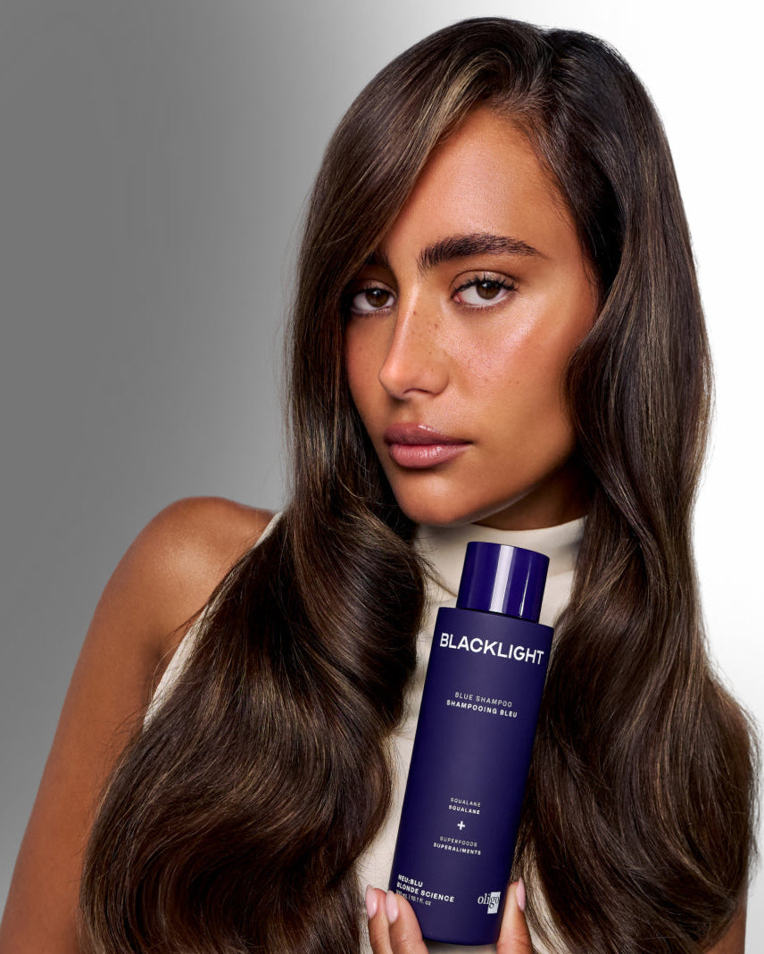 A woman with long, straight brown hair holds a bottle of BLACKLIGHT Hair Enhancing Hair Conditioner against a neutral gray studio background.