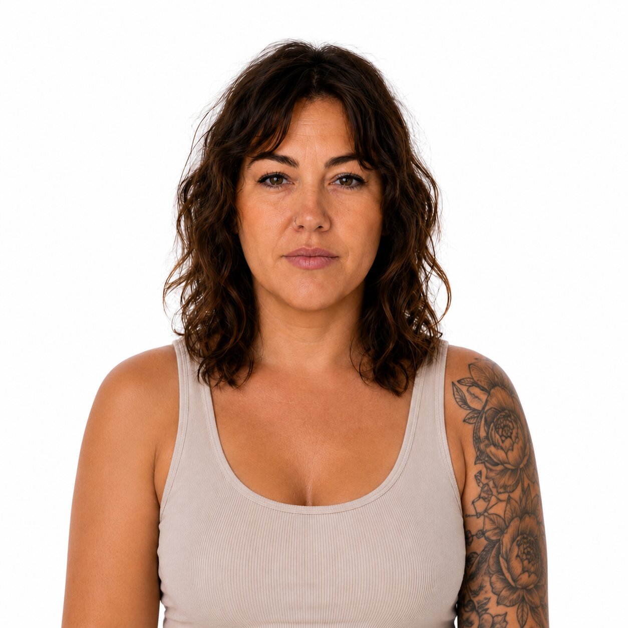A woman with shoulder-length wavy brown hair and a tattooed left arm wearing a light beige ribbed tank top, photographed against a solid white background.