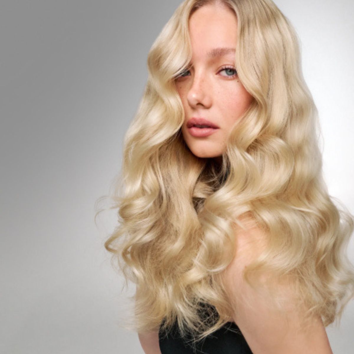 A woman with long, wavy platinum blonde hair wearing a black top, shown from the chest up against a gray gradient background.